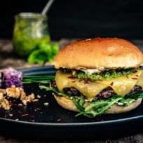 Ein Gourmet-Pesto-Walnuss-Burger mit geschmolzenem Vintage Cheddar, Pesto, Rucola und einem Brioche-Brötchen liegt auf einem schwarzen Teller. Walnüsse, essbare Blüten und Pesto in einem Glas stehen daneben auf einer rustikalen Holzoberfläche.