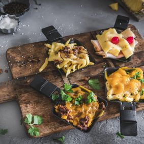 Vier kleine Raclette-Pfännchen auf einem Holzbrett. Oben links: Käsespätzle mit geschmolzenem Käse und Zwiebeln. Oben rechts: Toast Hawaii mit zusammengerolltem Schinken, Ananas und geschmolzenem Käse, garniert mit zwei Kirschtomaten. Unten rechts: Mac and Cheese mit cremiger Käsesauce und frischen Kräutern. Unten links: Chili con Carne mit Käse überbacken, garniert mit Kräutern. Auf dem Brett liegen verstreut Kräuterblätter, Salz und Pfefferkörner. Links oben im Bild ein Stück Baguette, rechts oben ein Messer