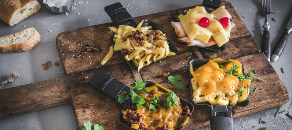 Vier kleine Raclette-Pfännchen auf einem Holzbrett. Oben links: Käsespätzle mit geschmolzenem Käse und Zwiebeln. Oben rechts: Toast Hawaii mit zusammengerolltem Schinken, Ananas und geschmolzenem Käse, garniert mit zwei Kirschtomaten. Unten rechts: Mac and Cheese mit cremiger Käsesauce und frischen Kräutern. Unten links: Chili con Carne mit Käse überbacken, garniert mit Kräutern. Auf dem Brett liegen verstreut Kräuterblätter, Salz und Pfefferkörner. Links oben im Bild ein Stück Baguette, rechts oben ein Messer