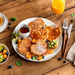 Herz-Pancakes mit Sirup, frischem Obst und einem Glas Orangensaft auf einem Holztisch. Besteck, eine kleine Serviette und eine Schale mit frischem Obst sind ebenfalls neben dem Hauptteller arrangiert.
