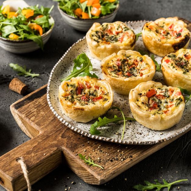 Ein Teller mit sechs mit Kräutern garnierten Mini-Quiches steht auf einem Holzschneidebrett, umgeben von frischem Salat, Paprika, einer Flasche Vinaigrette und verstreutem Rucola – perfekt für verschiedenste Rezeptkategorien.