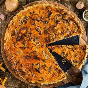 Eine frisch gebackene goldene Quiche mit Pilzen liegt auf einem Holzbrett, ein Stück davon wird serviert. Zutaten wie Pilze, Zwiebeln, Knoblauch und Gewürze sind auf einem rustikalen Holztisch verteilt.