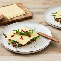 Zwei Teller mit belegten Brötchen mit Schnittkäse, Rucola und sonnengetrockneten Tomaten auf Körnerbrot stehen auf einem Holztisch. Auf einem Teller liegt ein Messer, das die Vielfalt von mild bis würzig präsentiert. Käsescheiben und Butter warten auf einem Brett daneben.