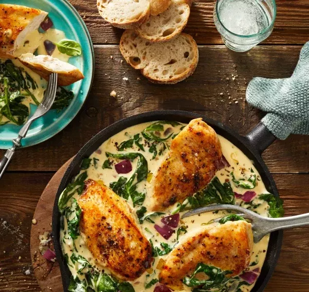 Eine Pfanne mit drei Stücken gewürztem, gekochtem Hühnchen auf einer cremigen Frischkäse-Spinat- und Rotzwiebelsauce steht auf einem Holztisch neben geschnittenem Brot, einem Teller mit einer Portion des Gerichts, einem Glas Wasser und Knoblauchzehen.