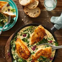 Eine Pfanne mit drei Stücken gewürztem, gekochtem Hühnchen auf einer cremigen Frischkäse-Spinat- und Rotzwiebelsauce steht auf einem Holztisch neben geschnittenem Brot, einem Teller mit einer Portion des Gerichts, einem Glas Wasser und Knoblauchzehen.