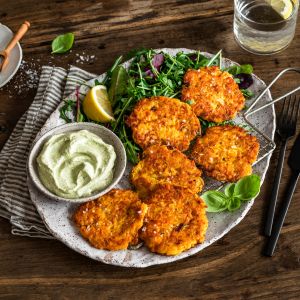 Ein Teller mit fünf goldenen Karottenpuffern auf Blattgemüse, einer Schale Kräuterdip, Zitronenspalten und einem Glas Wasser mit Zitrone auf einem rustikalen Holztisch. Besteck und eine gestreifte Serviette liegen neben dem Teller.