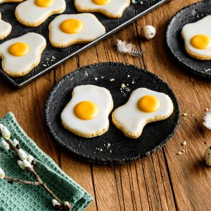 Spiegelei Kekse, dekorativ wie echte Spiegeleier gestaltet, liegen auf schwarzen Tellern und einer Tablette. Federn, Wachteleier und ein grünes Serviettendekor machen sie zu perfekten Osterplätzchen auf dem Holztisch.