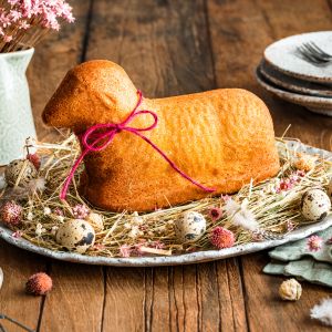 Ein lammförmiger Kuchen mit einer rosa Schleife wird auf einer Platte mit Heu, Wachteleiern und getrockneten Blumen präsentiert. Teller, Besteck und eine Vase mit rosa Blumen stehen auf einem rustikalen Holztisch.