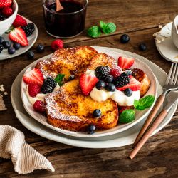 Ein Teller mit French Toast, garniert mit Sahne, Puderzucker und frischen Beeren wie Erdbeeren, Blaubeeren und Brombeeren, steht auf einem rustikalen Holztisch, daneben stehen Sirup und Besteck.