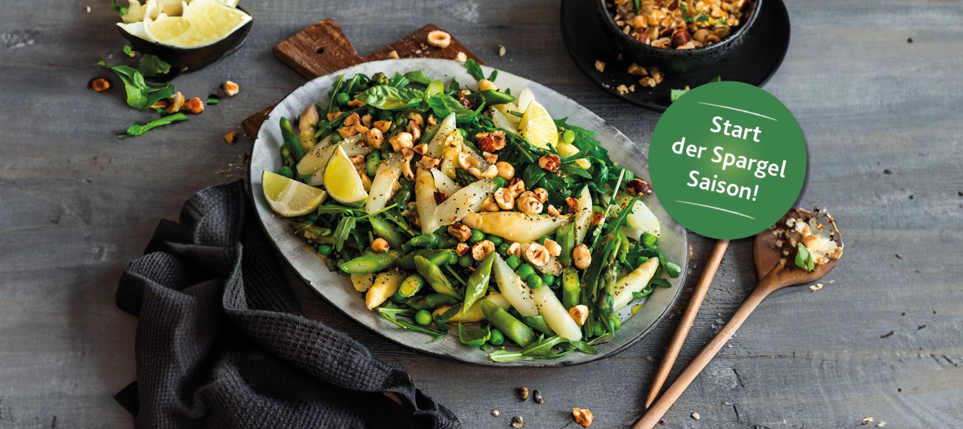 Eine Platte mit frischem Spargelsalat und Blattgemüse, garniert mit gerösteten Haselnüssen und Zitronenspalten. Daneben liegen ein Holzlöffel und eine Serviette. Auf dem Bild ist ein grüner Aufkleber mit der Aufschrift „Start der Spargelsaison!“ angebracht.