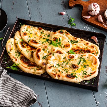 Auf einem blauen Holztisch steht ein Tablett mit vier Stücken Naan-Brot, garniert mit Kräutern, neben Petersilie, Knoblauch, einer Schüssel mit geschmolzener Butter, einer gestreiften Serviette und einem Holzbrett mit Salz.