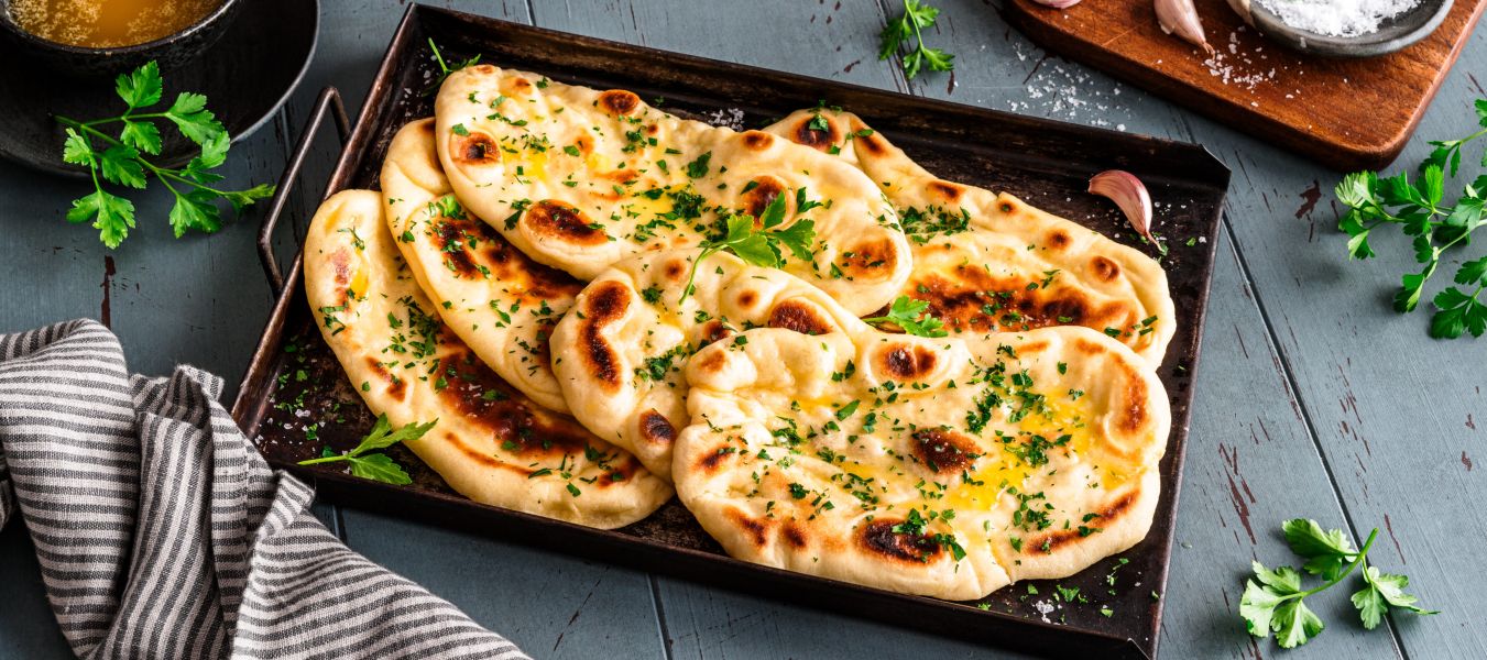 Auf einem blauen Holztisch steht ein Tablett mit vier Stücken Naan-Brot, garniert mit Kräutern, neben Petersilie, Knoblauch, einer Schüssel mit geschmolzener Butter, einer gestreiften Serviette und einem Holzbrett mit Salz.