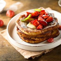 Ein Stapel Brioche-French-Toast mit Erdbeerscheiben, Puderzucker und einem Minzblatt liegt auf einem weißen Teller mit Erdbeeren und einer kleinen Schüssel Sirup in der Nähe auf einem Holztisch.