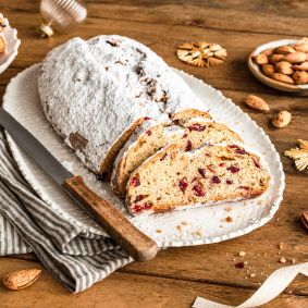 Ein mit Puderzucker bestäubter Christstollen, angeschnitten auf einer weißen Platte mit einem Messer. Die Scheiben geben den Blick auf Früchte und Nüsse frei. Umgeben von Mandeln, Keksen, einer Serviette und festlicher Weihnachtsdekoration auf einem Holztisch.