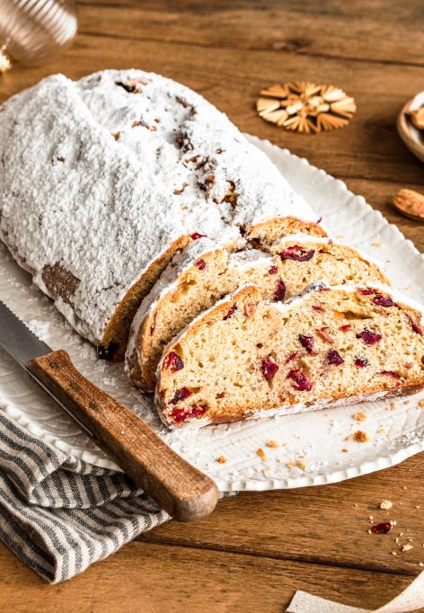 Ein mit Puderzucker bestäubter Christstollen, angeschnitten auf einer weißen Platte mit einem Messer. Die Scheiben geben den Blick auf Früchte und Nüsse frei. Umgeben von Mandeln, Keksen, einer Serviette und festlicher Weihnachtsdekoration auf einem Holztisch.