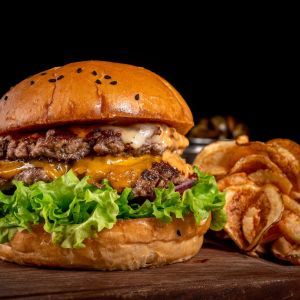 Ein saftiger Smashed Double Cheeseburger mit Salat, Cheddar-Käse und einem getoasteten Brötchen liegt auf einem Holzbrett neben knusprigen Kartoffelchips, im Hintergrund steht eine kleine Schale mit Essiggurken.