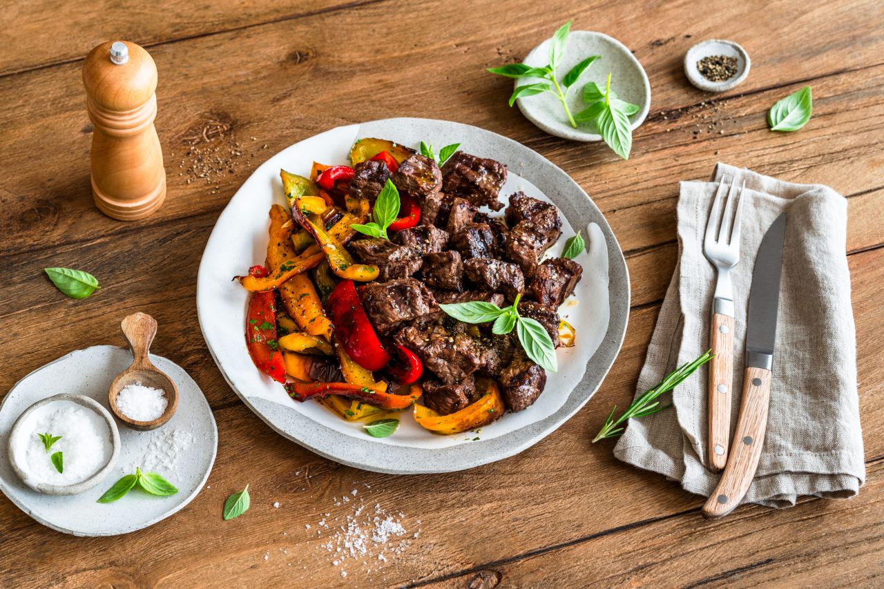 Ein Teller mit gegrillten Rindersteak-Häppchen mit buntem Grillgemüse und frischem Basilikum, serviert auf einem rustikalen Holztisch mit Salz, Pfeffer und in eine Serviette gewickeltem Besteck. Perfekt für Fans kreativer Steak-Rezepte.