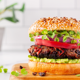 Nahaufnahme eines Gourmet-Burgers mit Sesambrötchen. Der Burger ist belegt mit Microgreens, roten Zwiebelscheiben, einer dicken Tomatenscheibe, einem gegrillten Patty mit Röstaromen und roten Paprikastückchen sowie einer Schicht Avocado-Creme auf dem unteren Brötchen. Im unscharfen Hintergrund ist etwas Grünpflanze in einem weißen Topf zu sehen.
