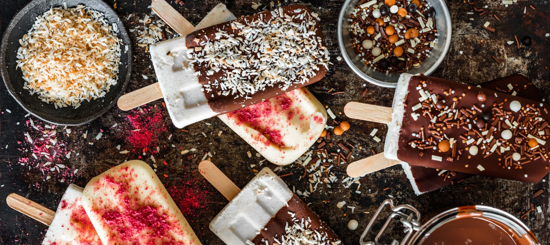 Verschiedene Schokoladen- und Vanilleeis am Stiel auf einer rustikalen Oberfläche, garniert mit Streuseln, Kokosflocken und rosa Pulver, umgeben von Schüsseln mit Toppings und geschmolzener Schokolade.