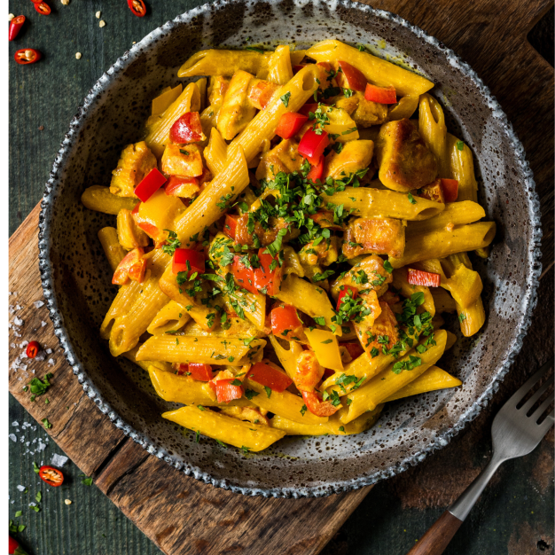 Eine Schüssel Penne-Nudeln mit Hühnchen, roten und gelben Paprikaschoten und frischen Kräutern steht auf einem Holzbrett, auf dem Chilischeiben verstreut liegen – eine köstliche Ergänzung für Ihre Rezeptkategorien-Sammlung.
