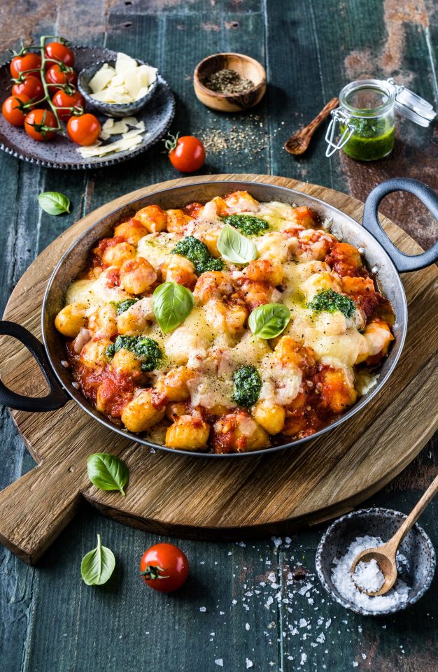 Eine runde Pfanne gefüllt mit gebackenen Gnocchi in Tomatensoße, geschmolzenem Käse und Pesto, garniert mit Basilikumblättern. Frische Tomaten, Parmesanscheiben, Salz und Pfeffer liegen auf einem rustikalen Holztisch daneben.