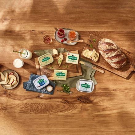 Ein Holztisch mit geschnittenem Brot, Arla Havarti-Käsepackungen, Käseecken, Apfelscheiben, einem Glas Milch, Butter, Kräutern und kleinen Schüsseln mit Marmelade.