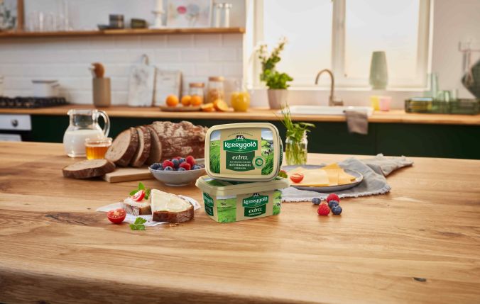 Zwei Becher mit irischer Butter Kerrygold Extra stehen auf einer hölzernen Küchentheke neben Brotscheiben, Beeren, einem Krug Milch und einem Teller Toast mit Butter in einer hellen und gemütlichen Küchenumgebung.