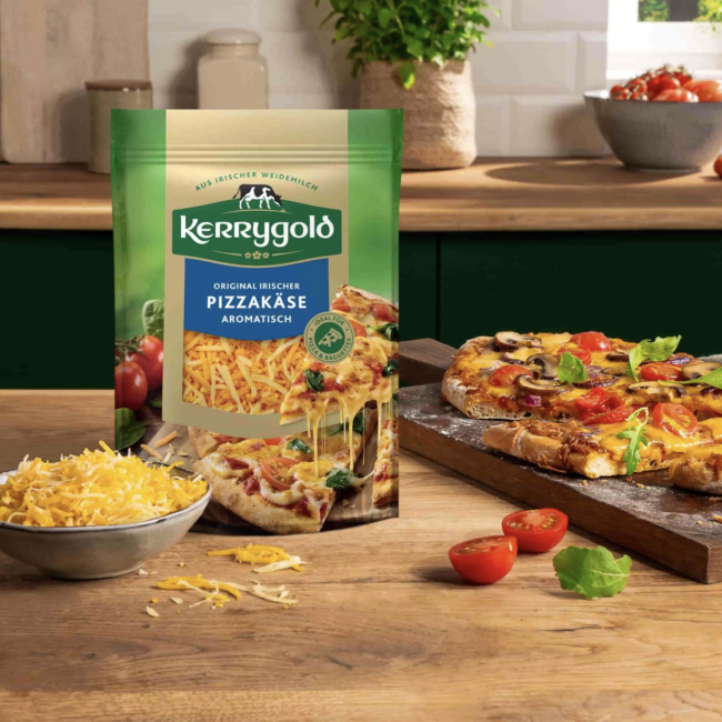 Eine Tüte geriebener Kerrygold-Pizzakäse liegt auf der Küchentheke neben einer Schüssel mit geriebenem Käse, frischen Tomaten, Basilikumblättern und einer geschnittenen selbstgemachten Pizza auf einem Schneidebrett. Sonnenlicht fällt durch ein Fenster herein.
