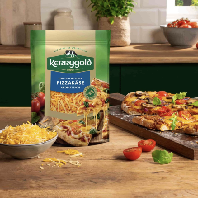Eine Tüte geriebener Kerrygold-Pizzakäse liegt auf der Küchentheke neben einer Schüssel mit geriebenem Käse, frischen Tomaten, Basilikumblättern und einer geschnittenen selbstgemachten Pizza auf einem Schneidebrett. Sonnenlicht fällt durch ein Fenster herein.