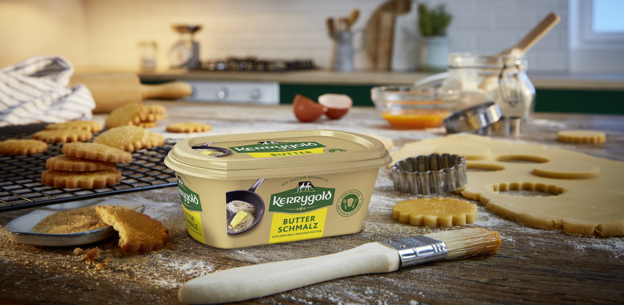 Ein Becher Kerrygold Butterschmalz steht auf einer Küchentheke, umgeben von Backzutaten, Keksteig, Backutensilien, einem Pinsel und frisch gebackenen Keksen, die auf einem Rost abkühlen.