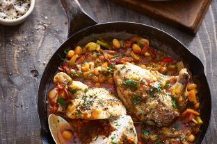 Eine Pfanne mit gebratenem Hähnchen, weißen Bohnen und Gemüse in einer Tomatensoße, garniert mit Kräutern. Daneben steht eine Schüssel Grünkohlsalat mit einem Stück Käse und einem Schälchen gesalzenem groben Salz.