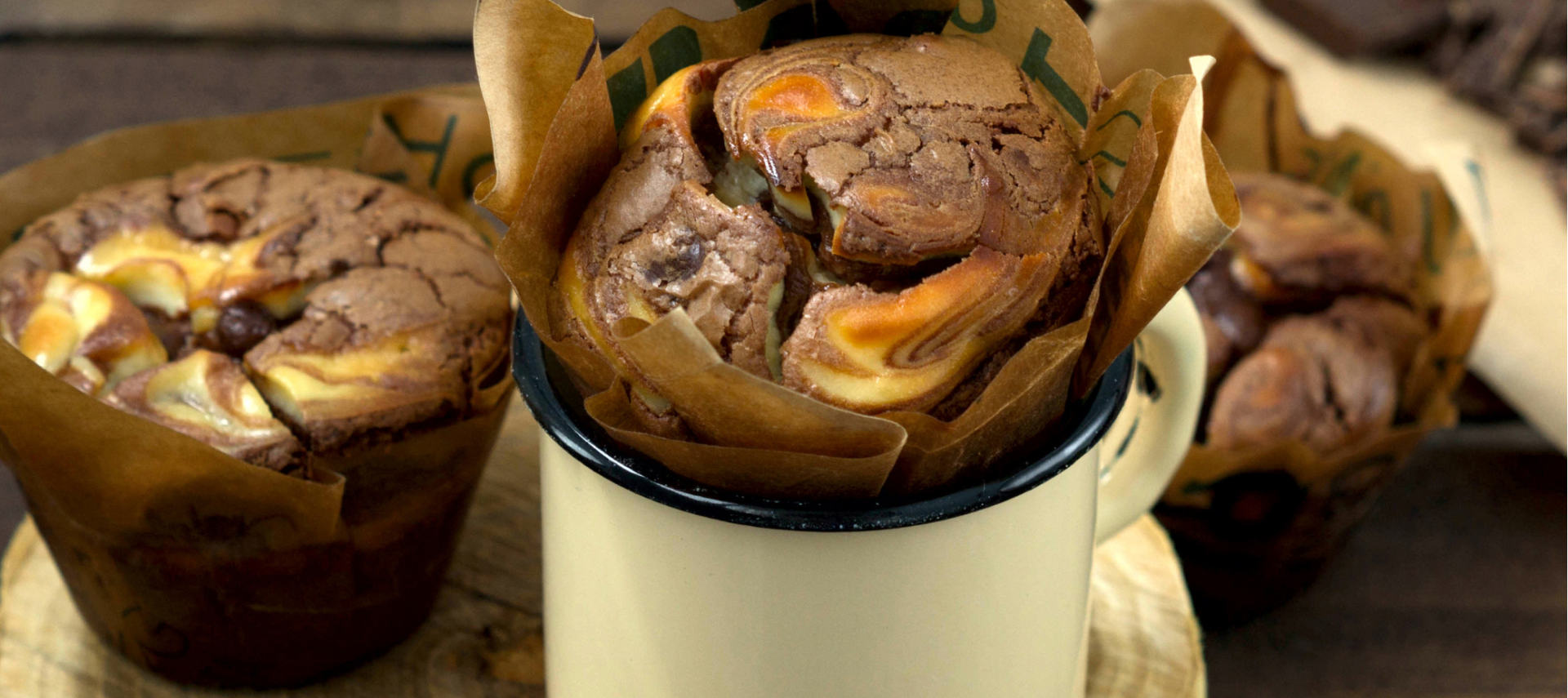 Drei Käsekuchen-Muffins mit Frischkäse-Spiral, einer in einer cremefarbenen Tasse auf einer Holzscheibe, zwei daneben auf einer rustikalen Holzoberfläche. Schokoladenstücke verschwimmen sanft im Hintergrund und heben diese unwiderstehlichen Schoko-Käsekuchen-Leckereien hervor.