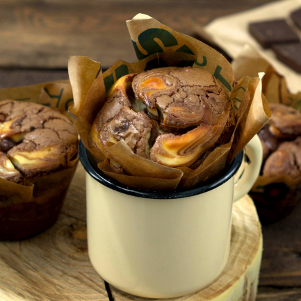 Drei Käsekuchen-Muffins mit Frischkäse-Spiral, einer in einer cremefarbenen Tasse auf einer Holzscheibe, zwei daneben auf einer rustikalen Holzoberfläche. Schokoladenstücke verschwimmen sanft im Hintergrund und heben diese unwiderstehlichen Schoko-Käsekuchen-Leckereien hervor.