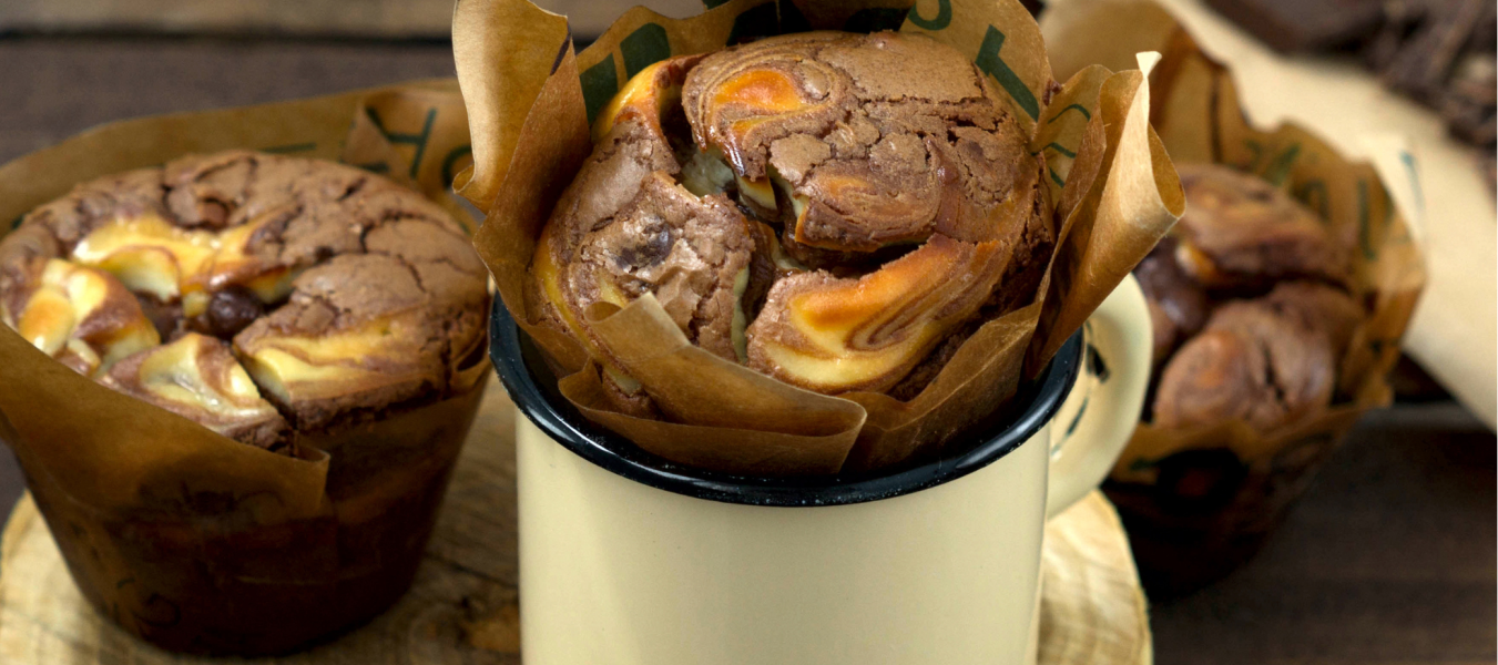 Drei Käsekuchen-Muffins mit Frischkäse-Spiral, einer in einer cremefarbenen Tasse auf einer Holzscheibe, zwei daneben auf einer rustikalen Holzoberfläche. Schokoladenstücke verschwimmen sanft im Hintergrund und heben diese unwiderstehlichen Schoko-Käsekuchen-Leckereien hervor.