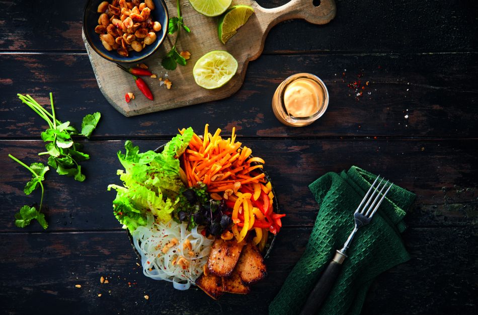 Bunte vietnamesische Nudelschüssel mit gegrilltem Fleisch, frischem Salat, Karotten, Paprika, Kräutern und Erdnüssen neben einer Dip-Sauce aus irischer Weidemilch, Limettenscheiben und Utensilien auf einem rustikalen Holztisch.