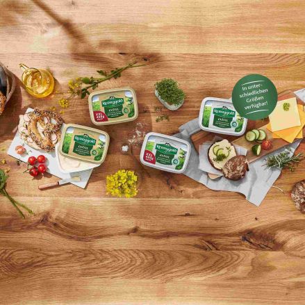 Ein Holztisch mit vier Wannen mit deutscher Streichfette, Brot, Brezeln, Käsescheiben, Kräutern, Kirschtomaten, Honig und einem Messer, angerichtet für eine rustikale Mahlzeit, mit einem grünen Produktkategorie-Etikett mit deutschem Text.