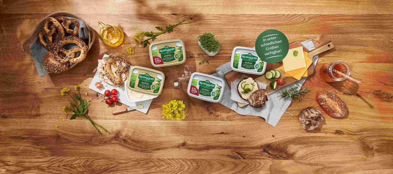 Ein Holztisch mit vier Wannen mit deutscher Streichfette, Brot, Brezeln, Käsescheiben, Kräutern, Kirschtomaten, Honig und einem Messer, angerichtet für eine rustikale Mahlzeit, mit einem grünen Produktkategorie-Etikett mit deutschem Text.