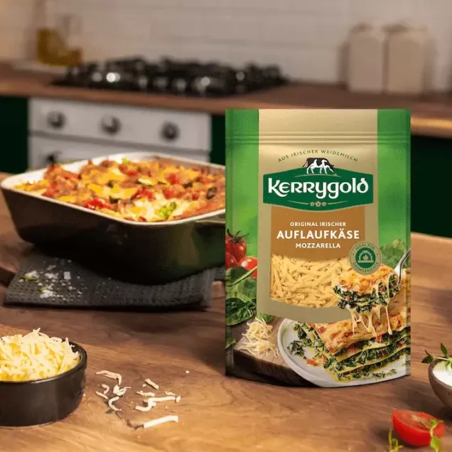Eine Packung Kerrygold Auflaufkäse Mozzarella liegt auf einer hölzernen Küchentheke, im Hintergrund stehen eine Schüssel mit geriebenem Auflaufkäse und ein gebackener Auflauf.
