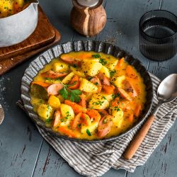 Ein rustikaler Teller mit Kartoffel-Möhren-Eintopf, bestehend aus Karottenscheiben und Wurst, ist mit Petersilie garniert und steht auf einer gestreiften Serviette neben einem Löffel, im Hintergrund stehen ein dampfender Topf mit Eintopf sowie Salz- und Pfefferstreuer.