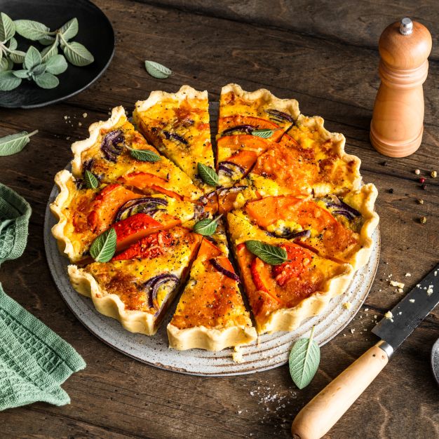 Eine in Scheiben geschnittene Kürbisquiche mit Tomaten und Kräutern liegt auf einem Teller, umgeben von einem Messer, Salz, einer Pfeffermühle, einem grünen Handtuch und einer schwarzen Schale mit frischen Salbeiblättern auf einem rustikalen Holztisch.