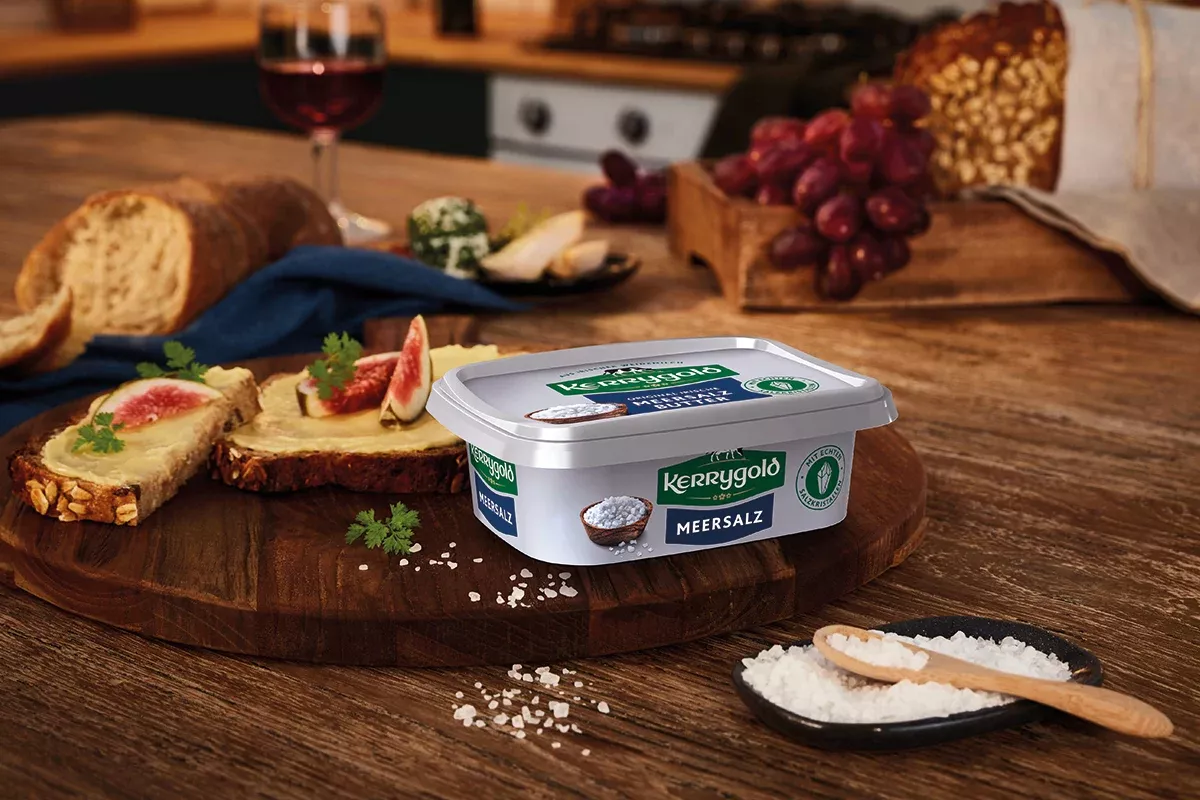 Ein Becher Kerrygold Meersalzbutter steht auf einem Holzbrett, umgeben von Brotscheiben mit Butter und Feigen. Eine Schüssel Salz, ein Laib Brot, Trauben, Käse und ein Glas Rotwein runden die einladende Szene ab.