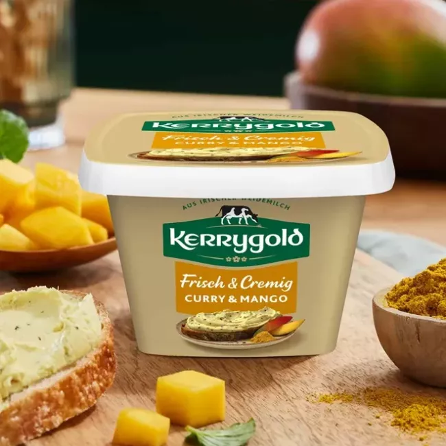 Ein Behälter mit Kerrygold Frisch & Cremig Curry & Mango-Aufstrich steht auf einem Holzbrett mit gewürfelter Mango, einer Schüssel Currypulver und einer Scheibe Brot mit dem cremigen Mango & Curry-Aufstrich und Kräutern. Im Hintergrund sind Mangos zu sehen.