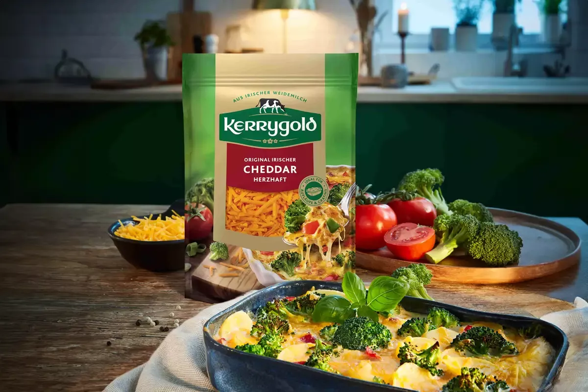 Eine Tüte herzhafter Kerrygold Cheddar Reibekäse steht auf einem Holztisch neben einer Schüssel mit Brokkoli-Auflauf, geriebenem Käse, frischem Brokkoli, Tomaten und Basilikum in einer gemütlichen Küchenumgebung.