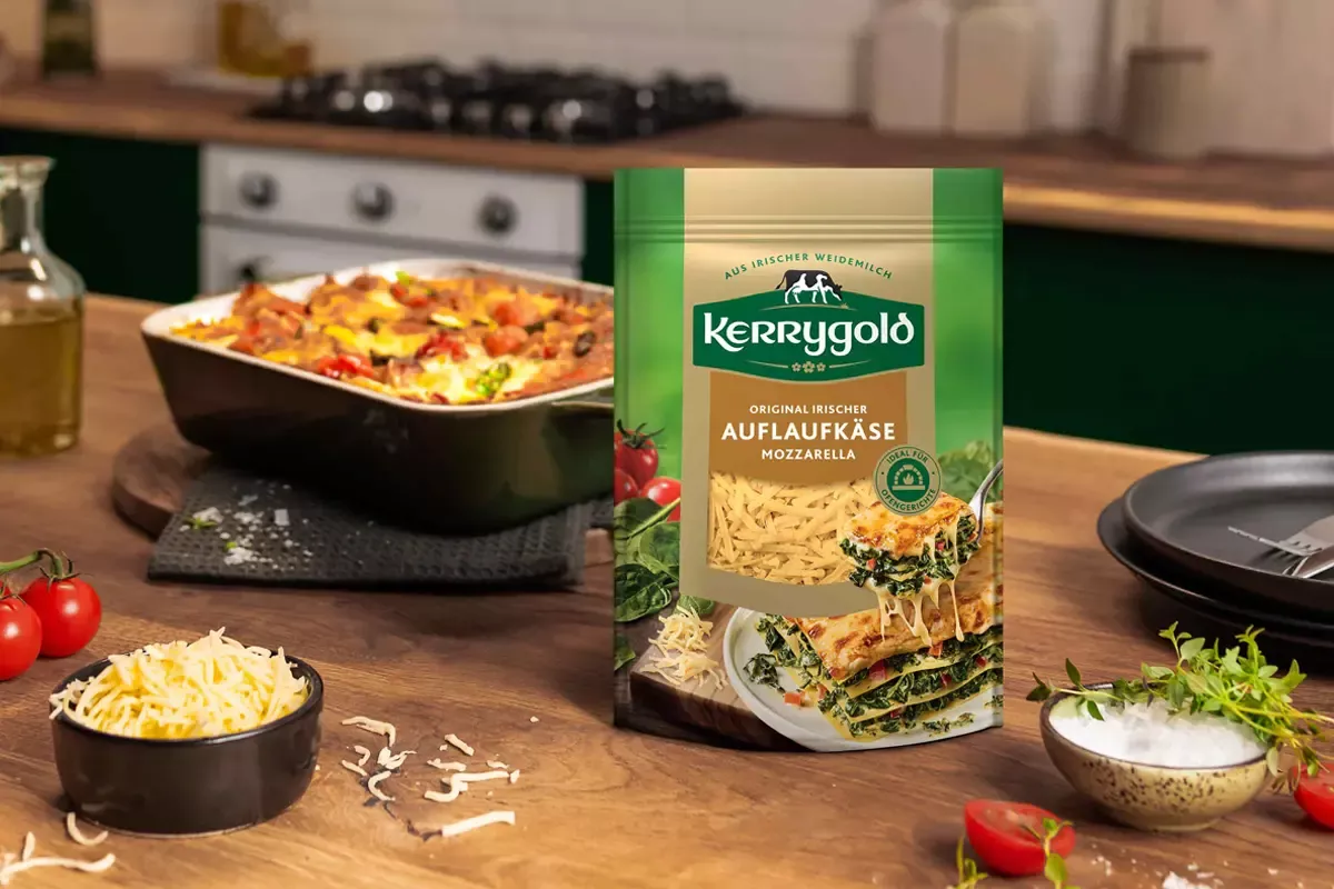 Eine Packung geriebener Mozzarella von Kerrygold, auf Deutsch als Auflaufkäse bekannt, liegt auf einer Küchentheke neben einer Schüssel mit Mozzarella, Tomaten, Salz, Besteck und im Hintergrund einer Auflaufform.