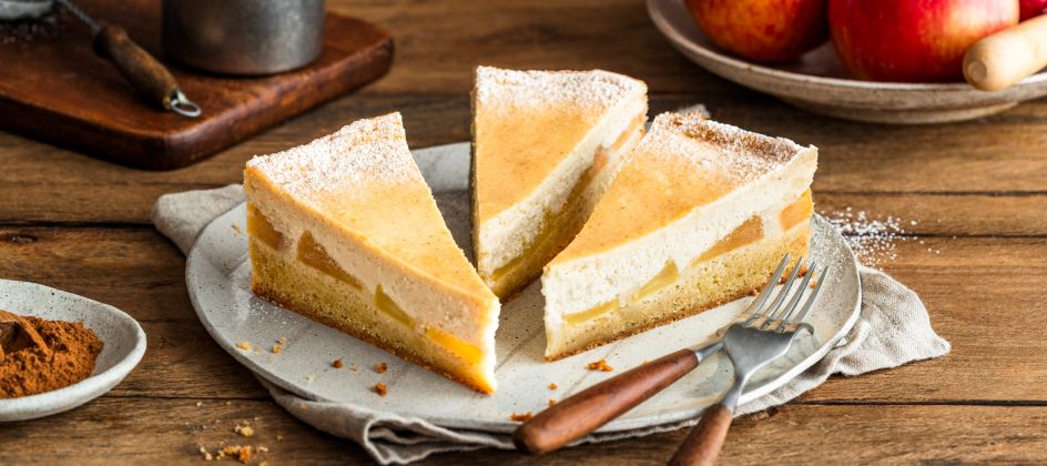 Drei Stücke Apfelkäsekuchen mit einer zarten Käsekuchenschicht liegen mit einer Gabel auf einem Teller auf einem Holztisch. Im Hintergrund sind frische Äpfel in einer Schüssel und Backutensilien zu sehen; der Kuchen ist leicht mit Puderzucker bestäubt.