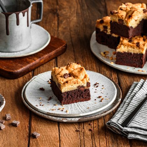 Auf einem Teller liegt ein quadratisches Stück Chocolate Chip Cookie Brownie, auf einem anderen Teller sind weitere Brownies gestapelt. Auf einem Holztisch stehen eine gestreifte Serviette mit einer schwarzen Gabel, eine Tasse mit Schokoladentropfen und eine kleine Schüssel mit Schokoladenstückchen.