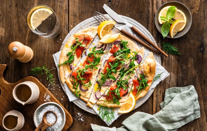 Ein Lachsflammkuchen mit Räucherlachs, Rucola, roten Zwiebeln und Kräutern liegt angeschnitten auf einem runden Teller. Zitronenspalten, frische Kräuter, ein Glas Wasser und kleine Schälchen sind auf einem rustikalen Holztisch angerichtet.