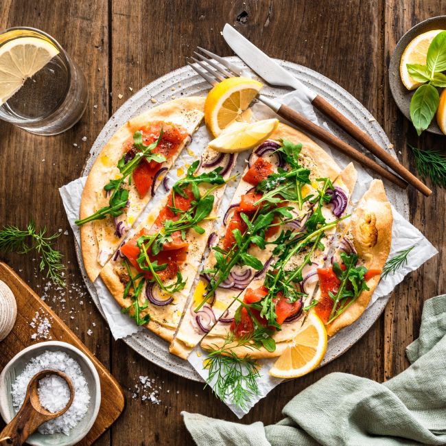 Ein Lachsflammkuchen mit Räucherlachs, Rucola, roten Zwiebeln und Kräutern liegt angeschnitten auf einem runden Teller. Zitronenspalten, frische Kräuter, ein Glas Wasser und kleine Schälchen sind auf einem rustikalen Holztisch angerichtet.