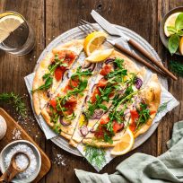 Ein Lachsflammkuchen mit Räucherlachs, Rucola, roten Zwiebeln und Kräutern liegt angeschnitten auf einem runden Teller. Zitronenspalten, frische Kräuter, ein Glas Wasser und kleine Schälchen sind auf einem rustikalen Holztisch angerichtet.