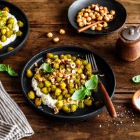 Zwei schwarze Teller mit Gnocchi sind mit kräftigem Pesto, cremigem Ricotta, gehackten Haselnüssen und frischem Basilikum garniert und stehen auf einem rustikalen Holztisch, umgeben von einer Schale mit Haselnüssen, einer Salzschale, einer Pfeffermühle und einer gestreiften Serviette.